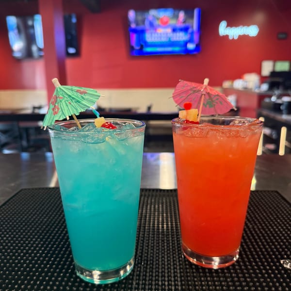 Two colorful cocktails, blue and red, with umbrellas and fruit garnishes on a bar counter.