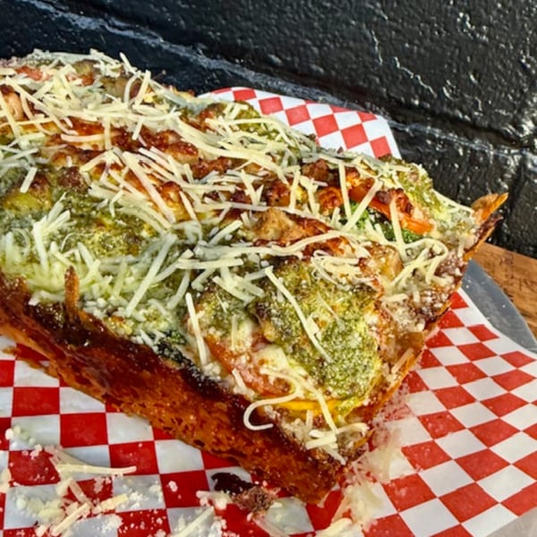 Detroit-style pizza with pesto, cheese, and other toppings on red and white checkered paper.