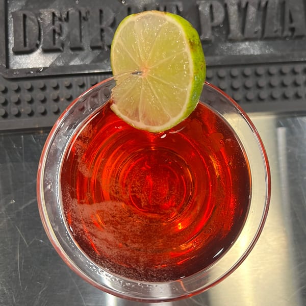 Top-down view of a red cocktail with a lime slice, on a bar mat with "DETROIT PIZZA" visible.